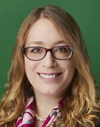 A person with long curly hair and glasses smiles at the camera against a green background. They are wearing a pink patterned scarf and pearl necklace.